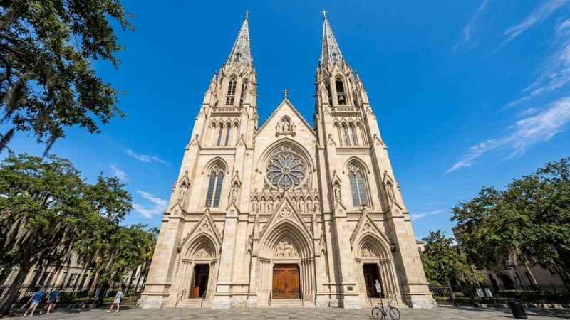Exterior facade and intricate twin spires of the Cathedral Basilica of St. John the Baptist in Savannah against a blue sky.