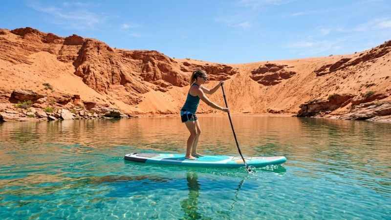 Paddleboarding on the clear blue water of Sand Hollow State Park reservoir with red sand dunes behind it.