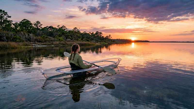 Clear kayak floating on calm glassy waters in St. Joseph Bay reflecting a warm orange and pink sunset with an uncrowded natural shoreline.