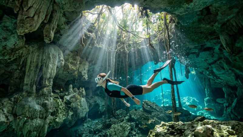 Swimmer exploring the crystal-clear fresh water and limestone rock formations inside Gran Cenote near Tulum.
