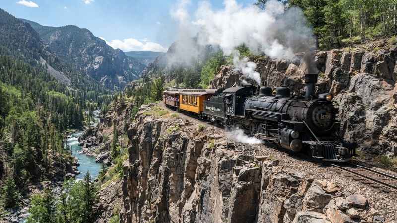 A vintage steam train travels along a steep rock cliff inside a narrow river canyon.